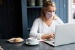 © Mint Images - Woman wearing face mask sitting alone at a cafe table with a laptop, working remotely.