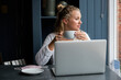 © Mint Images - Young blond woman sitting alone at a cafe table with a laptop computer, working remotely.