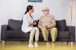 © LIGHTFIELD STUDIOS - geriatric nurse talking to senior man while sitting on sofa at home on blurred foreground