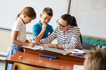 Wall Mural - In the classroom's cozy setting, the caring female teacher diligently checks her elementary school students' homework, providing guidance and support to nurture their academic growth and success.