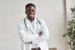 Happy african male doctor wearing white coat, stethoscope looking at camera standing with arms crossed in medical office. Smiling black man professional therapist physician posing at wor, portrait.