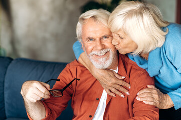 Happy elderly gray-haired couple hugging and sits on the sofa at home, wife kisses husband and husband looks at the camera and smiles