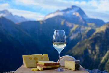 Tasty cheese and wine from Savoy region in France, beaufort, abondance, emmental, tomme and reblochon de savoie cheeses and glass of white wine served outdoor with Alpine mountains peaks on background