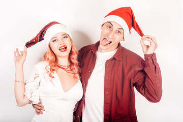 Couple on a white background in santa hats. The girl and the guy are smiling, new year, christmas. Woman in a beautiful white dress and pink hair, in accessories. Lovers grimace and have fun