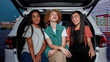 © Svitlana - A close up of three young commonly dressed women of different nationalities sitting in an opened car trunk outside on a parking site smiling and laughing looking into a camera