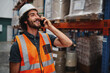 © StratfordProductions - Happy bearded warehouse manager in conversation on mobile phone wearing white helmet and safety vest standing looking away