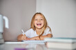 © Svitlana - Happy laughing little girl sitting at the white desk and doing writing exercises in book