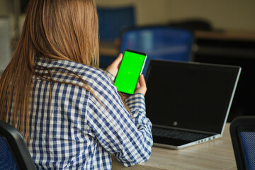 Wall Mural - girl with glasses holding a phone with a green screen sitting at a laptop.
