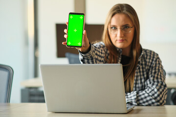 Wall Mural - girl with glasses holding a phone with a green screen sitting at a laptop.