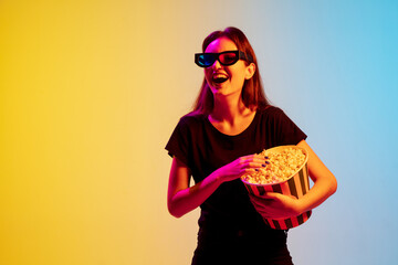 Wall Mural - Laughting. Young caucasian woman with bunch of popcorn on gradient blue-yellow studio background in neon. Concept of movie, human emotions, facial expression, sales, ad. Watching cinema in eyewear.