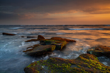 Wall Mural - Beautiful seascape. Beach with stones covered by seaweeds. Low tide. Composition of nature. Motion water. Cloudy sky with sunlight. Slow shutter speed. Mengening beach, Bali, Indonesia