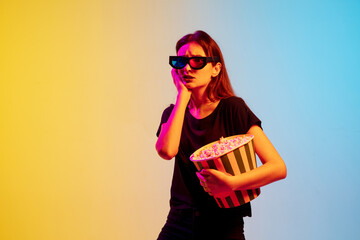 Wall Mural - Sad. Young caucasian woman with bunch of popcorn on gradient blue-yellow studio background in neon. Concept of movie, human emotions, facial expression, sales, ad. Watching cinema in eyewear.