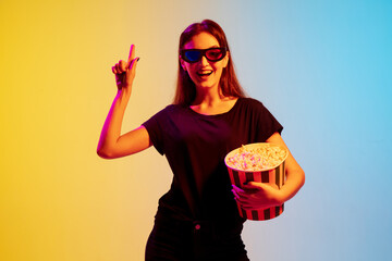 Wall Mural - Pointing. Young caucasian woman with bunch of popcorn on gradient blue-yellow studio background in neon. Concept of movie, human emotions, facial expression, sales, ad. Watching cinema in eyewear.