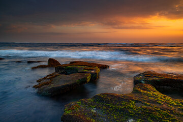 Wall Mural - Beautiful seascape. Beach with stones covered by seaweeds. Low tide. Composition of nature. Motion water. Cloudy sky with sunlight. Slow shutter speed. Mengening beach, Bali, Indonesia