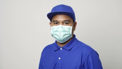 Wall Mural - Asian delivery man in blue uniform wearing protection mask and medical rubber gloves standing on isolated white background. 4k Resolution.