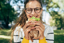 Closeup Portrait Of A Happy Student Female Has Lunch With A Healthy Sandwich Outdoors. A Blonde Young Woman In Glasses Takes A Rest To Eat Fast Food In The Park.