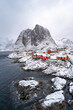 a small serene Norwegian fishing village on a shore in the north. The town, big mountain, and cliff covered with white fluffy snow in the beautiful winter under overcast sky. Waves break on the rocks.