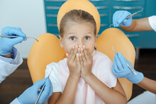Little Girl Afraid And Covers Her Mouth With Hands In Dentists Chair In Interior Of Modern Clinic