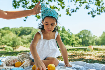 Wall Mural - Horizontal outdoor image of the cute little girl sitting on the picnic blanket play and counting the harvest of the lemons with family on a sunny day. Child spend time with family in the park.