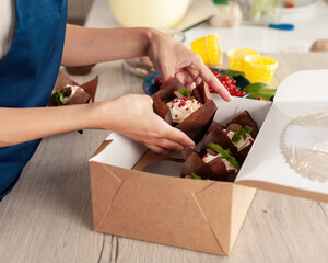 Closeup view of hands of female confectioner or baker in blue apron packing fresh decorated muffins with cream top to delivery craft box. Ready cupcakes with berries and mint in package.