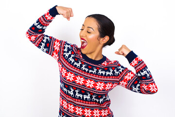 Wall Mural - Yes I am winner. Portrait of charming delighted and excited Young beautiful Arab woman wearing Christmas sweater raising up fist in triumph and victory smiling achieving success grinning from delight.