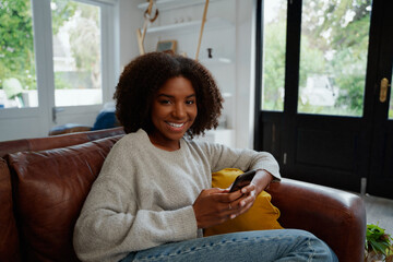 Wall Mural - Portrait of cheerful african woman holding mobile phone sitting on couch looking at camera