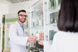 © LIGHTFIELD STUDIOS - happy pharmacist in eyeglasses and white coat giving bottle with pills to brunette colleague on blurred foreground
