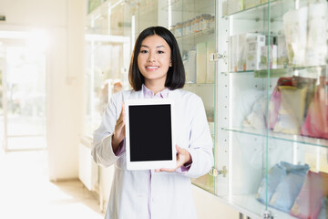 Wall Mural - cheerful asian pharmacist in white coat holding digital tablet with blank screen in drugstore