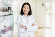 cheerful asian pharmacist in white coat standing with crossed arms in drugstore