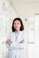 Wall Mural - brunette asian pharmacist in white coat standing with crossed arms in drugstore