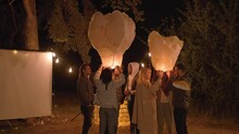 Sequence Of Shots Of Joyful Company Of Close Friends Preparing Chinese Lanterns For Flying Standing In Forest Decorated With String Lights In Middle Of Night