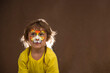 a child with a painted lynx mask thinks about it and looks up on a brown background. The hair of the little girl is highlighted from behind