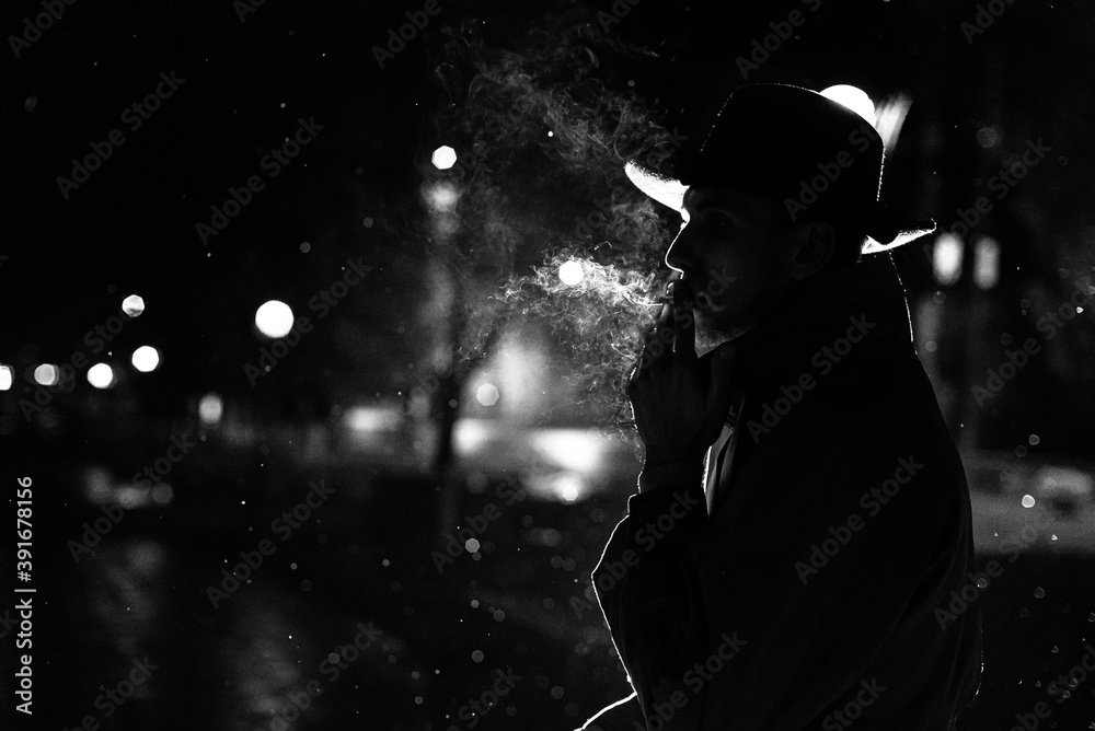dark silhouette of a man in a hat Smoking a cigarette in the rain on a  night street Stock Photo
