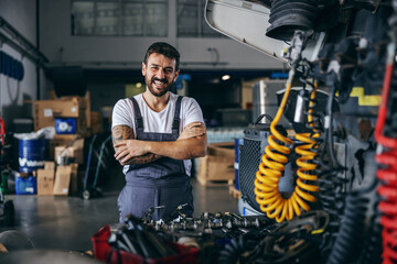 Smiling happy bearded tattooed worker in overalls standing next to truck with arms crossed.