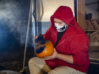 Wall Mural - Person in a red hoodie and a skull facemask shading a carved Halloween pumpkin