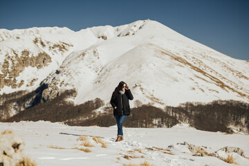 Wall Mural - Young woman doing winter hike in white snowy mountains. Female tourist walks on mountain, blue sky, trees and white mountains in background. 