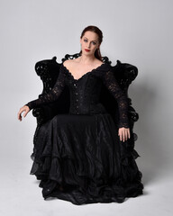 full length portrait of  woman wearing black gothic dress, sitting on a ornate black armchair. Seated pose, against a studio background.