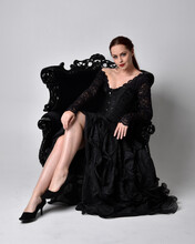 Full Length Portrait Of  Woman Wearing Black Gothic Dress, Sitting On A Ornate Black Armchair. Seated Pose, Against A Studio Background.