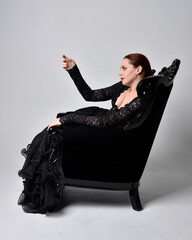 full length portrait of  woman wearing black gothic dress, sitting on a ornate black armchair. Seated pose, against a studio background.