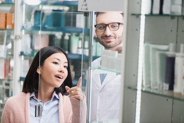 Wall Mural - curious asian customer pointing with finger near bearded pharmacist in drugstore