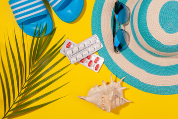 Wall Mural - Top view on white and red pills in blister pack, blue hat, blue flip-flops, sunglasses, seashell, palm leaf on yellow background. Concept of beach holiday, sea tour, warm sunny summer. Acclimatization