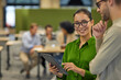 © Friends Stock - Two young coworkers, man and woman using digital tablet, and discussing something while standing in the coworking space with colleagues on the background