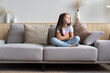 © opolja - Beautiful unhappy sad little 6-year old girl sitting cross legged on couch in living room, feeling bored and lonely, being home alone while her parents spending all day at work