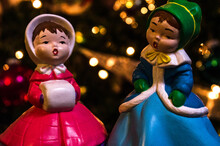 Christmas Scene Of Two Figurine Girls Singing In Front Of A Xmas Tree Of Green And Gold Bokah Lights.