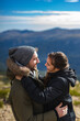 © Jota_Visual - Young couple in love and in winter clothes hug and smile in front of the mountain range in a sunny day
