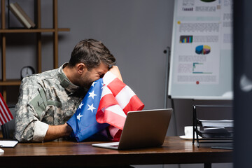 Wall Mural - military man in uniform crying and covering face with flag of america near laptop