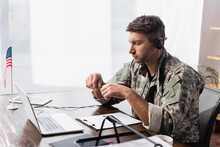 Patriotic Military Man In Headset Holding Pen And Looking At Laptop Near American Flag