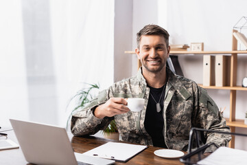 Wall Mural - cheerful military man in uniform holding cup of coffee near laptop on desk