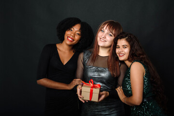 Wall Mural - Indoor portrait of three happy bestfriends sisters women, ready for holiday party, holding bright gifts and presents, wearing bright dresses on black background