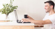 © Pablo - Retrato de un hombre adulto y apuesto caucásico sentado frente a su laptop y bebiendo una taza de café mientras la usa en un apartamento brillante y blanco con luz natural.